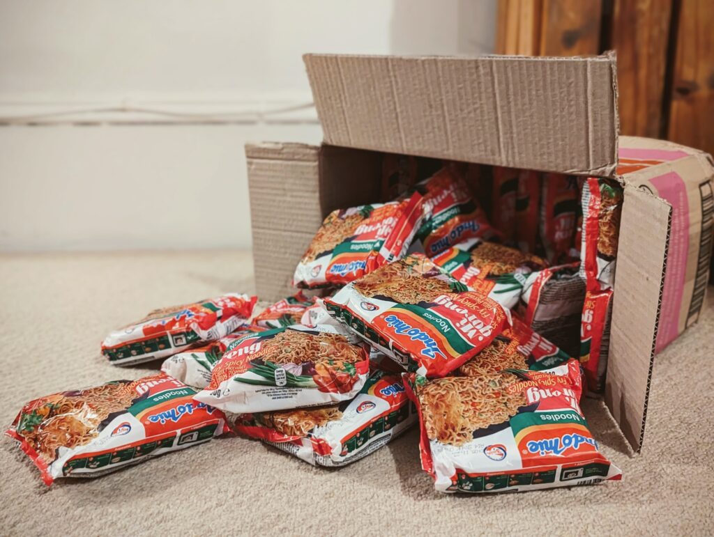 Indomie Mi Goreng instant noodle packets spilling out of an open cardboard box onto a carpeted floor.