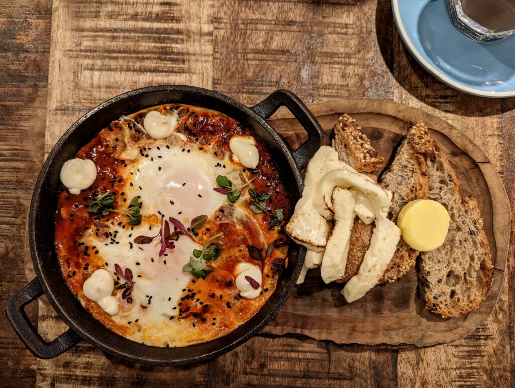 Shakshuka in a black skillet with eggs and herbs, served with grilled halloumi and seeded sourdough toast on a wooden board.