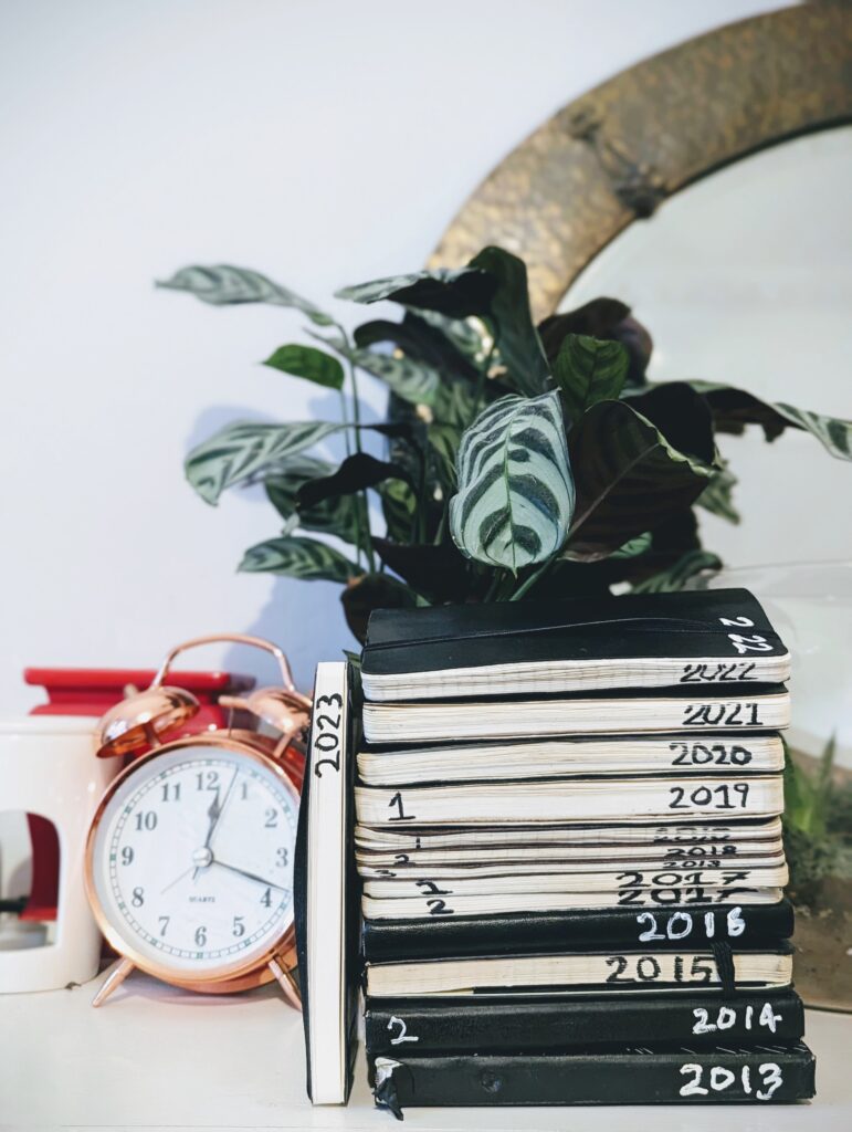 A stack of black notebooks hand-labeled 2013 to 2022 beside a 2023 journal, a copper alarm clock, and a patterned Calathea plant.