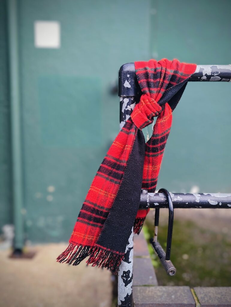 A red and black tartan scarf tied to a weathered black metal railing with peeling paint against a blurred green background.
