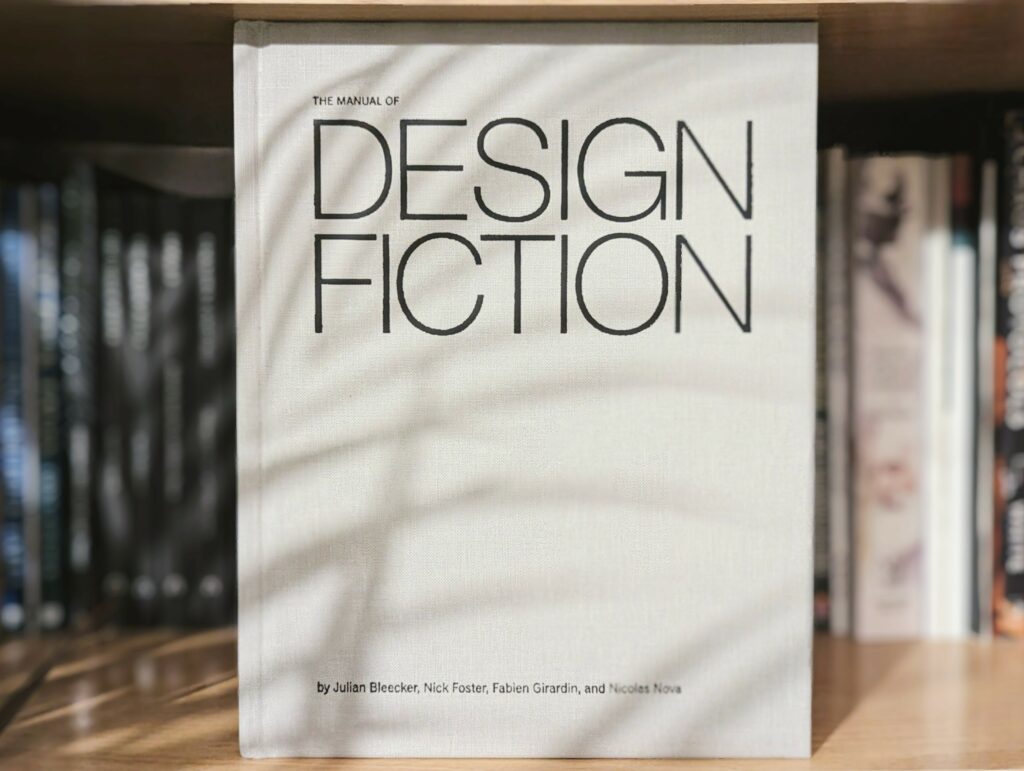 The Manual of Design Fiction book on a wooden shelf, its white cover dappled with long palm leaf shadows.