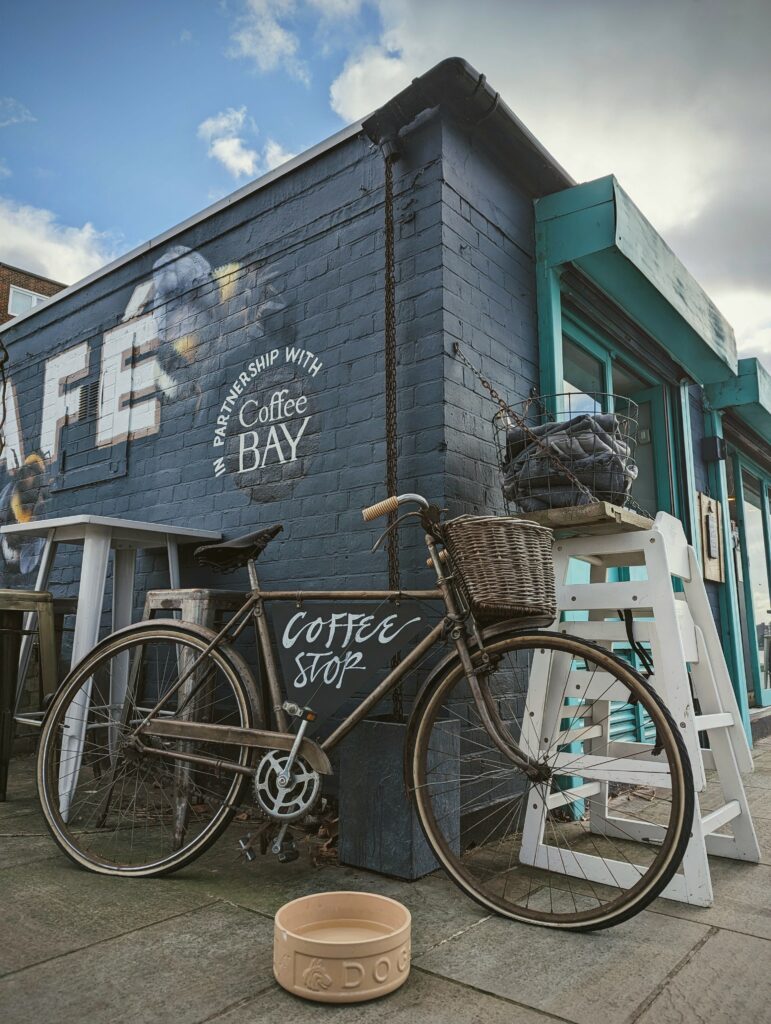 A vintage bicycle with a wicker basket and Coffee Stop sign parked outside a blue-grey brick cafe with a bumblebee mural and dog bowl.