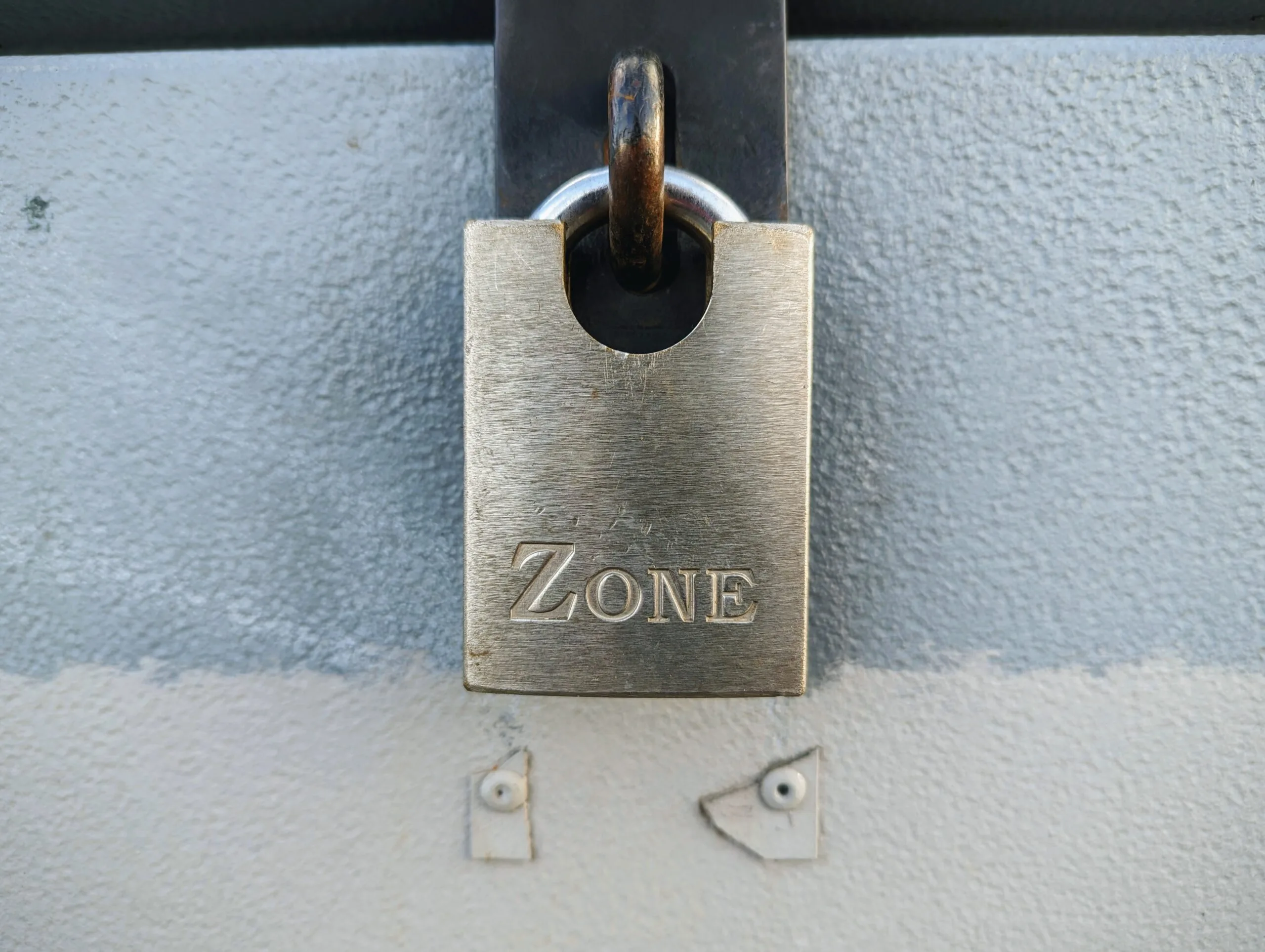A rectangular silver padlock with the word ZONE engraved on it, hanging from a black metal latch on a textured grey surface.