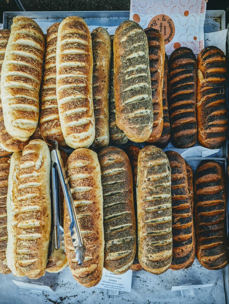 Rows of assorted artisanal bread loaves, from cheesy golden crusts to dark seeded varieties, on a market tray with metal serving tongs.