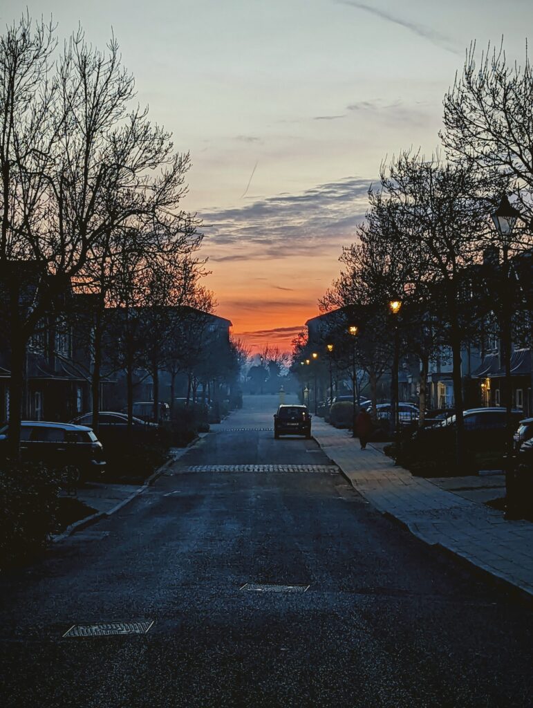 A residential street framed by bare trees leading towards a vibrant orange sunset, with streetlights glowing and parked cars in deep shadow.