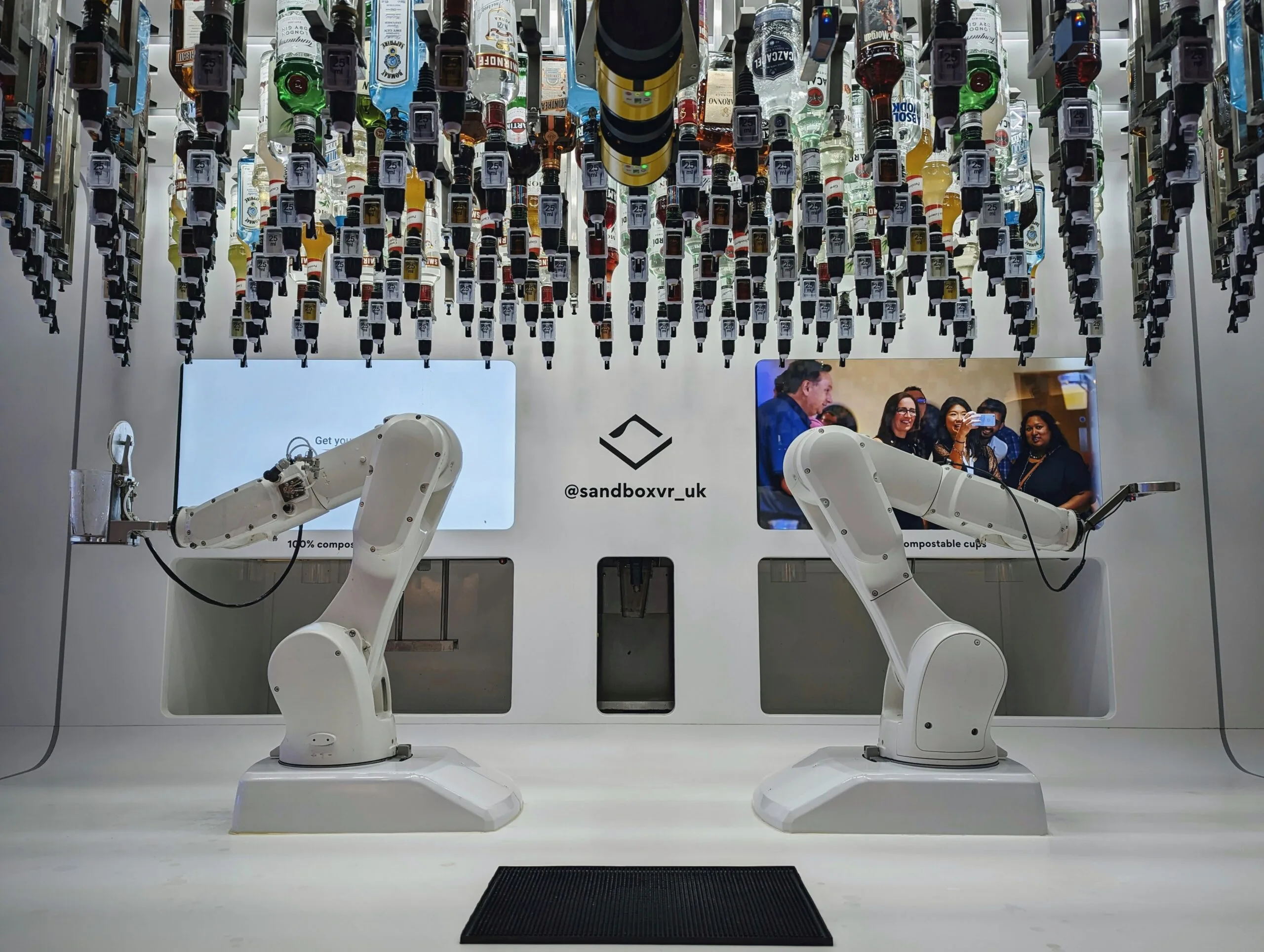 Two white robotic arms at a Sandbox VR UK automated bar, positioned beneath dozens of liquor bottles mounted upside down on the ceiling.