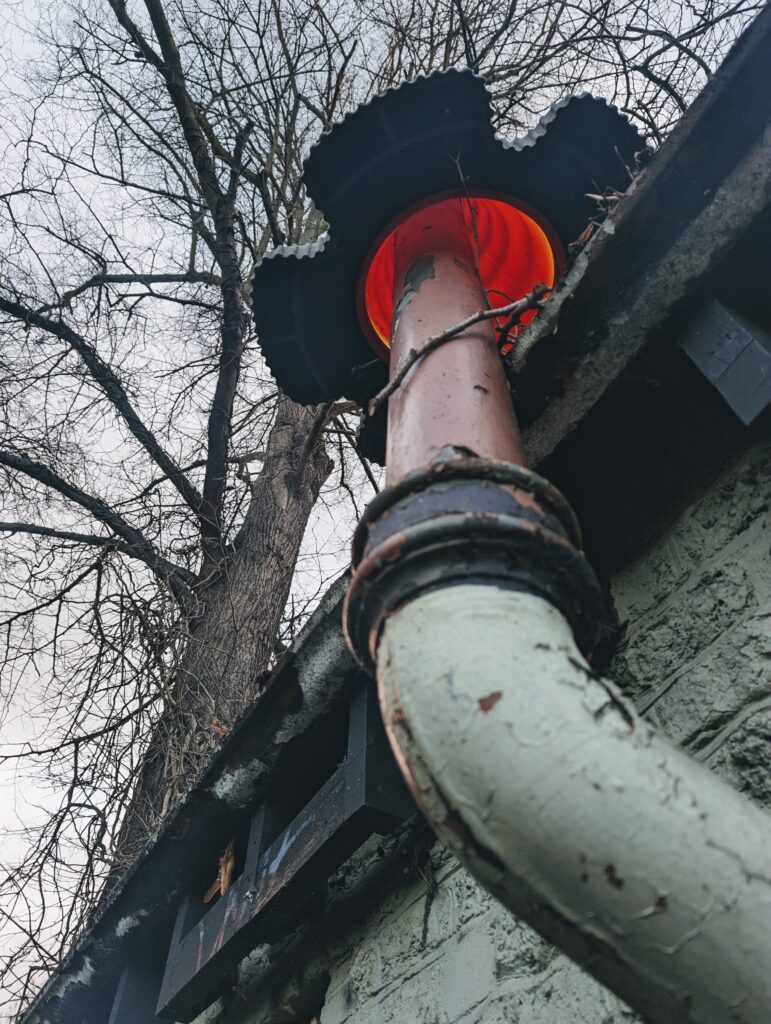 Weathered pipe on a building with a glowing orange interior, seen from below against bare winter trees and a grey sky.