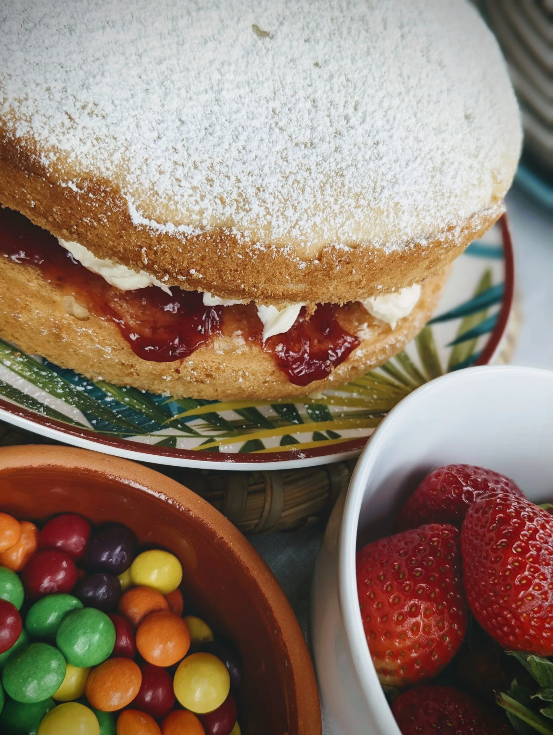 A Victoria sponge cake dusted with icing sugar and filled with jam and cream, next to bowls of colorful sweets and fresh strawberries.