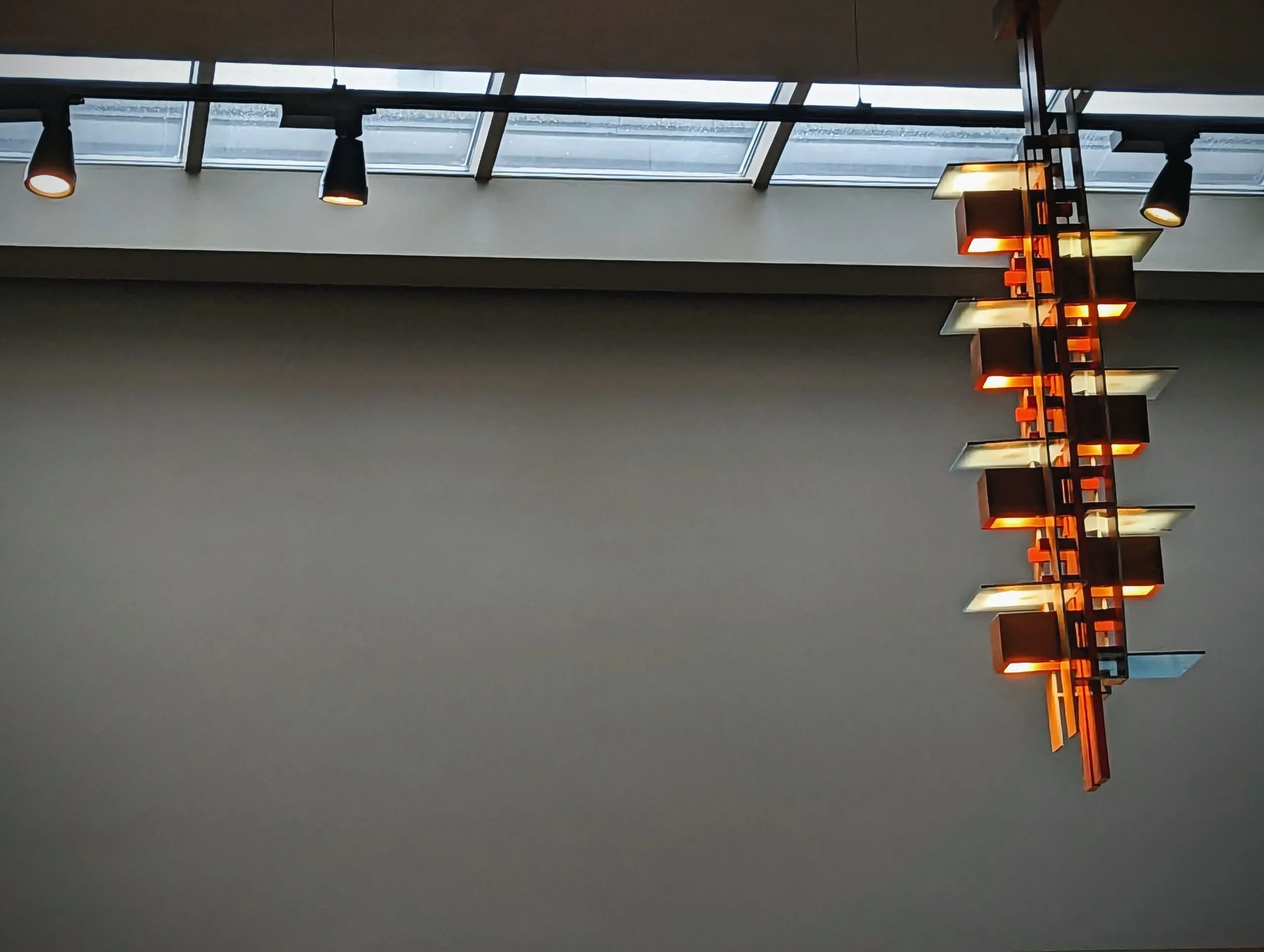 A geometric wooden pendant lamp with warm orange glowing cubes hanging against a plain grey wall under gallery track lighting and a skylight.