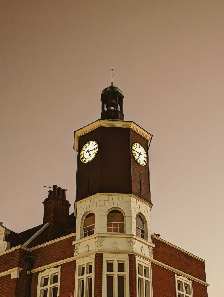 Illuminated clock faces on an octagonal tower above a red brick building against a hazy, warm-toned twilight sky.