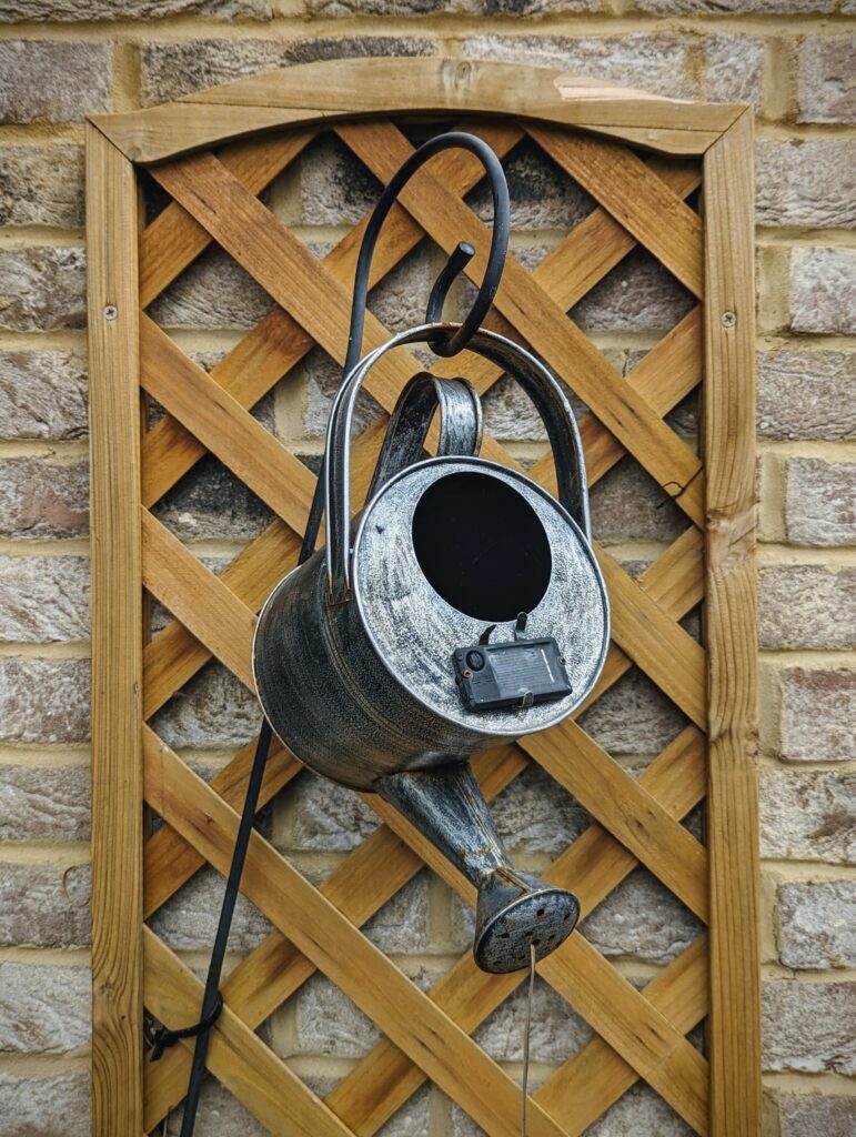 A weathered metal watering can solar light hanging from a black hook in front of a wooden garden trellis on a brick wall.