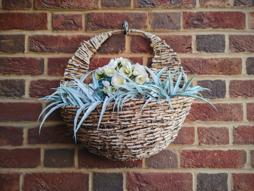 A rustic woven wall basket filled with white flowers and pale spiky leaves, hanging on a red brick wall.