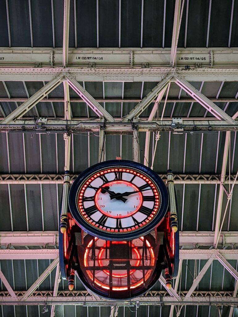 Station clock at Waterloo in London with a red neon inner ring and Roman numerals, suspended from the grey industrial rafters of a station roof.