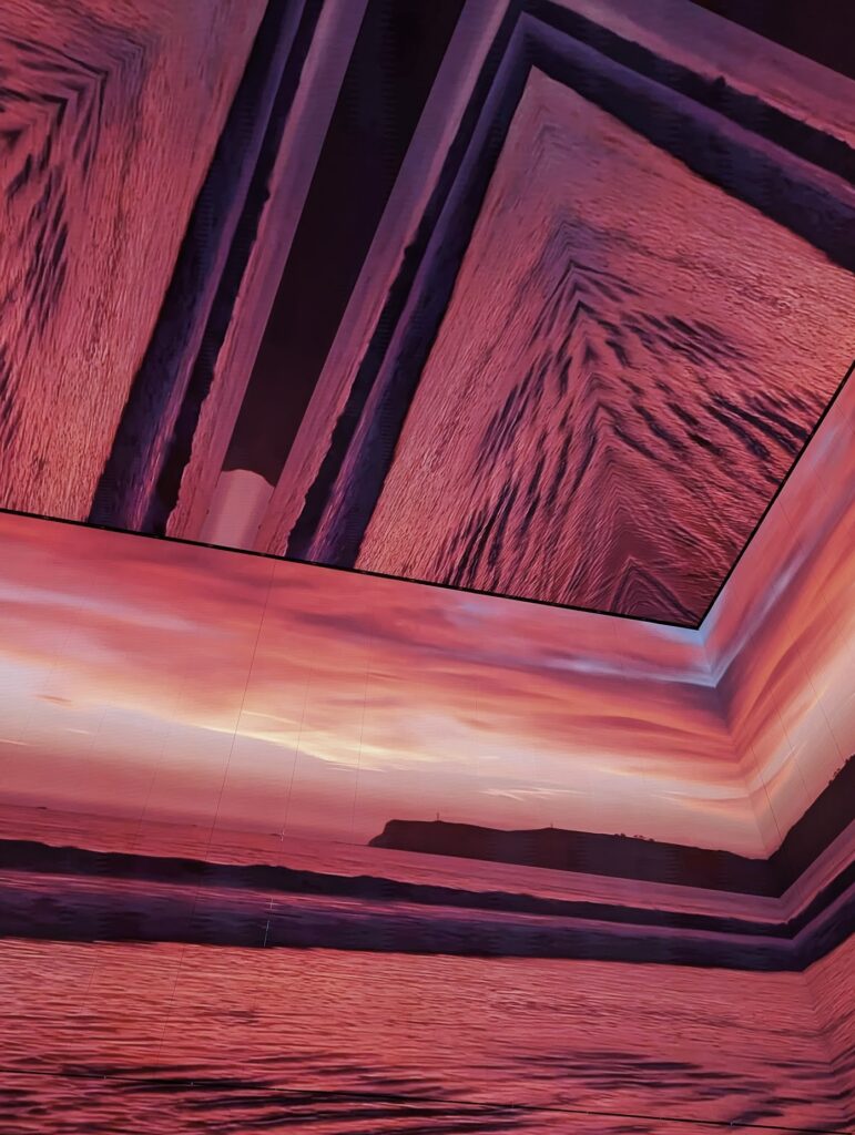 A digital projection of a pink sunset over the sea and a dark coastline, mirrored across the walls and ceiling panels of an immersive installation.