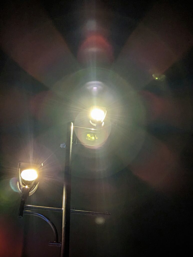 Two glowing street lamps on a pole at night, with dramatic circular lens flares and starburst light rays against a dark background.