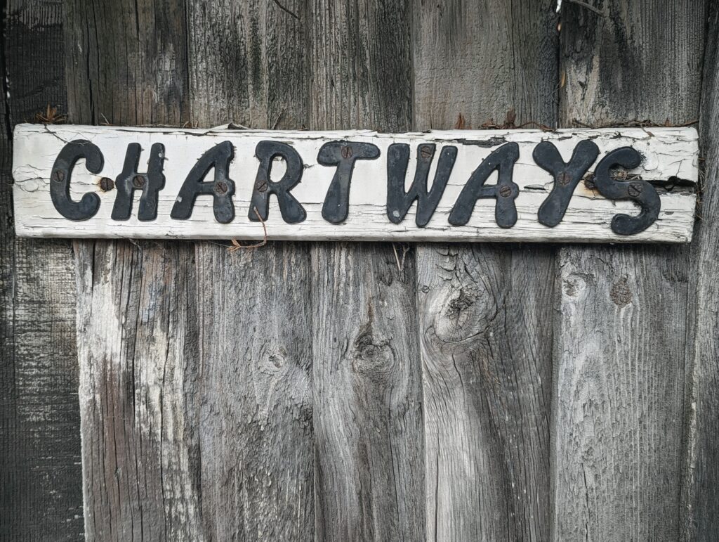Black metal letters spelling CHARTWAYS on a weathered white wooden sign, mounted against a rustic grey wood plank fence.