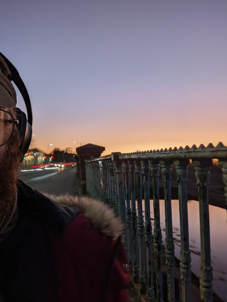 A bearded person wearing headphones on a bridge with ornate green railings during a purple and orange sunset.