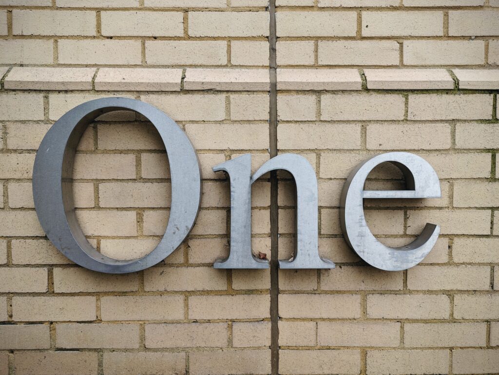 Large 3D brushed metal letters spelling the word One mounted on a light-coloured brick wall.