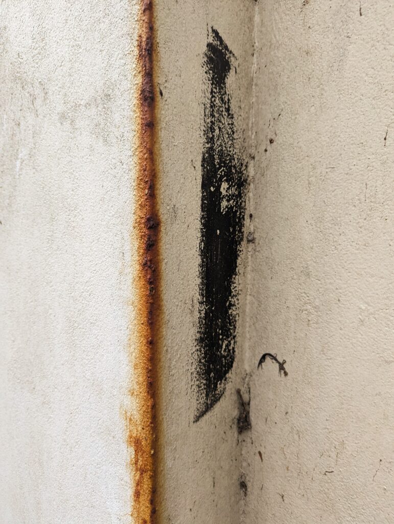 A vertical orange rust stain along the edge of a textured off-white wall, with a black paint smudge in the corner.