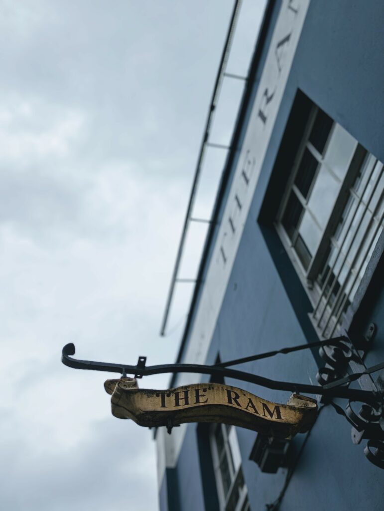 A scroll-style pub sign reading THE RAM hangs from an ornate iron bracket on a blue building facade under a grey overcast sky.