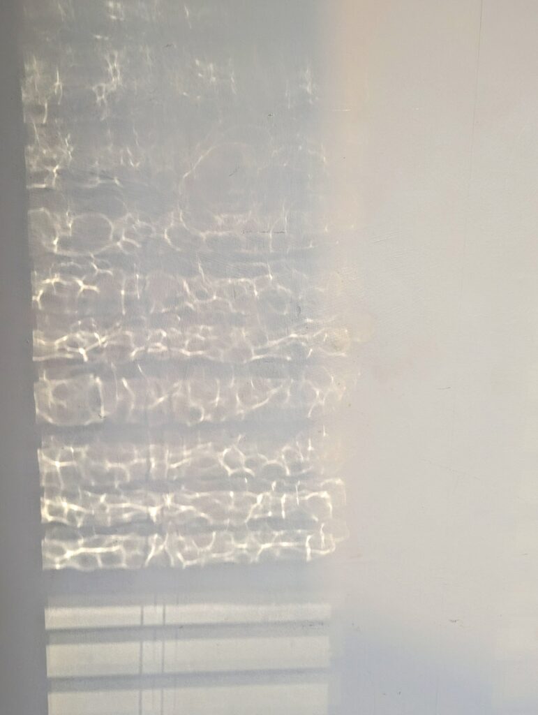 Shimmering caustic light patterns reflected from water onto a white wall, above horizontal shadows from window blinds.