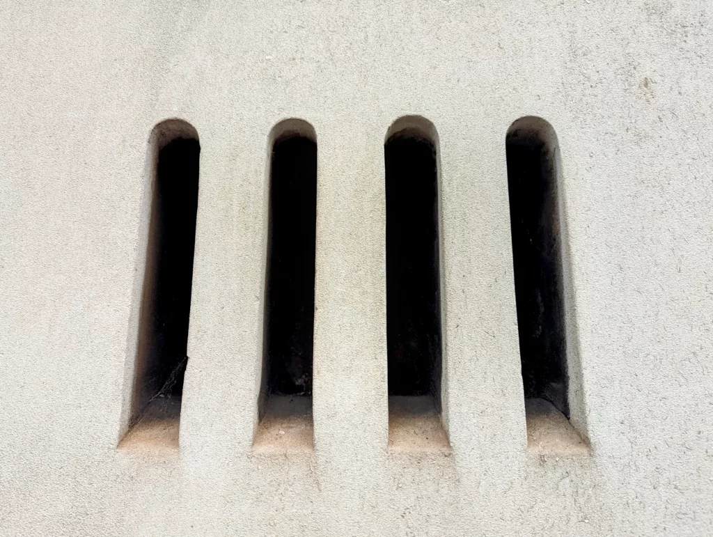 Four narrow, vertical arched vents in a light-colored textured wall, opening into dark shadows.