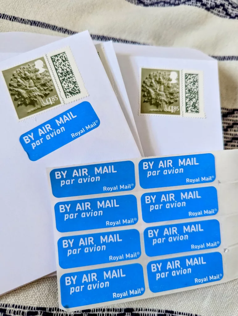 Two white envelopes with green Royal Mail oak tree stamps beside a sheet of blue air mail stickers on a patterned fabric surface.