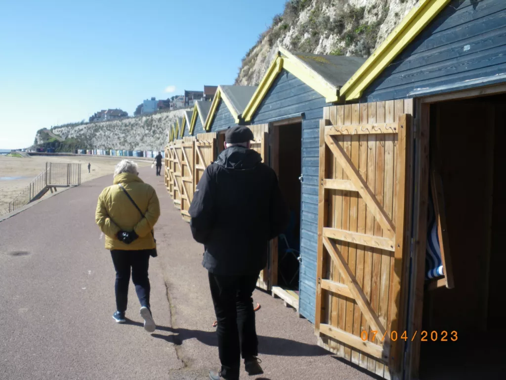 A woman in a yellow jacket and a man in black walking past blue beach huts on a coastal promenade beneath white cliffs.