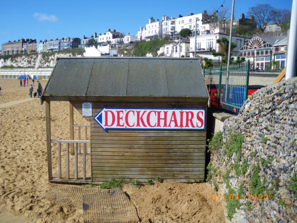 A wooden beach hut on sand with a large sign reading DECKCHAIRS inside a blue arrow. Coastal houses line the cliff in the background.