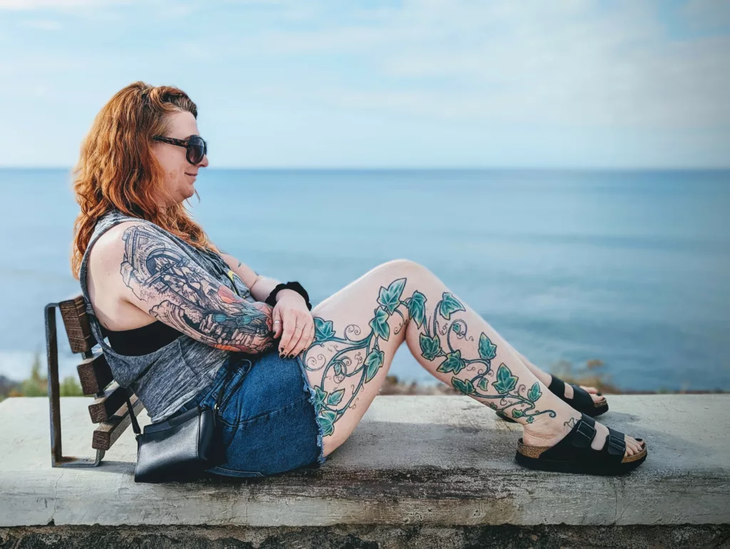 A woman with red hair and ivy leg tattoos sits on a stone ledge overlooking the sea, wearing sunglasses and denim shorts.