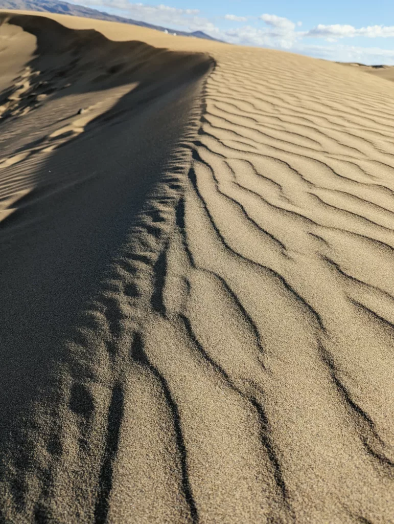 Wind-blown ripples on a golden sand dune with a sharp shadow line, under a blue sky with distant hills.