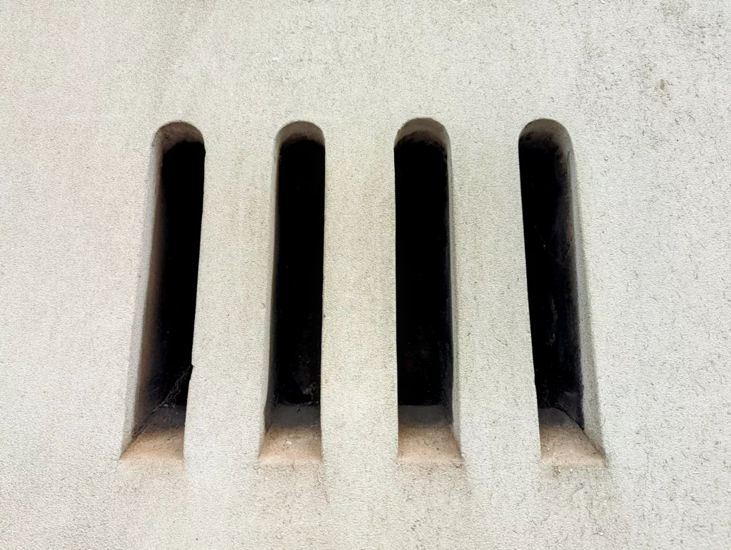 Four narrow, vertical arched slits with dark interiors set into a light-colored, textured wall.