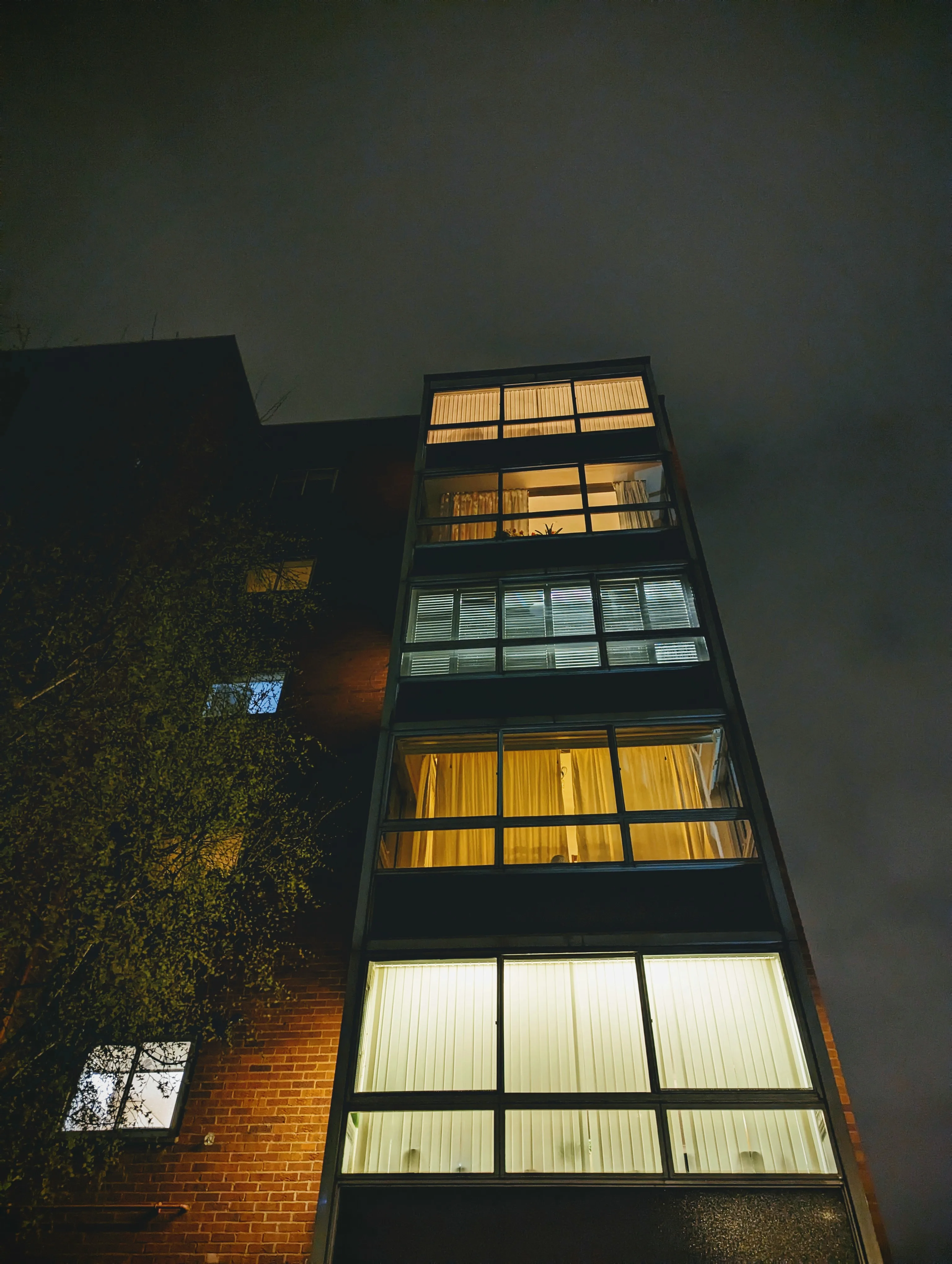 Upward view of a brick apartment building at night, with a vertical column of glowing windows and blinds against a dark, cloudy sky.