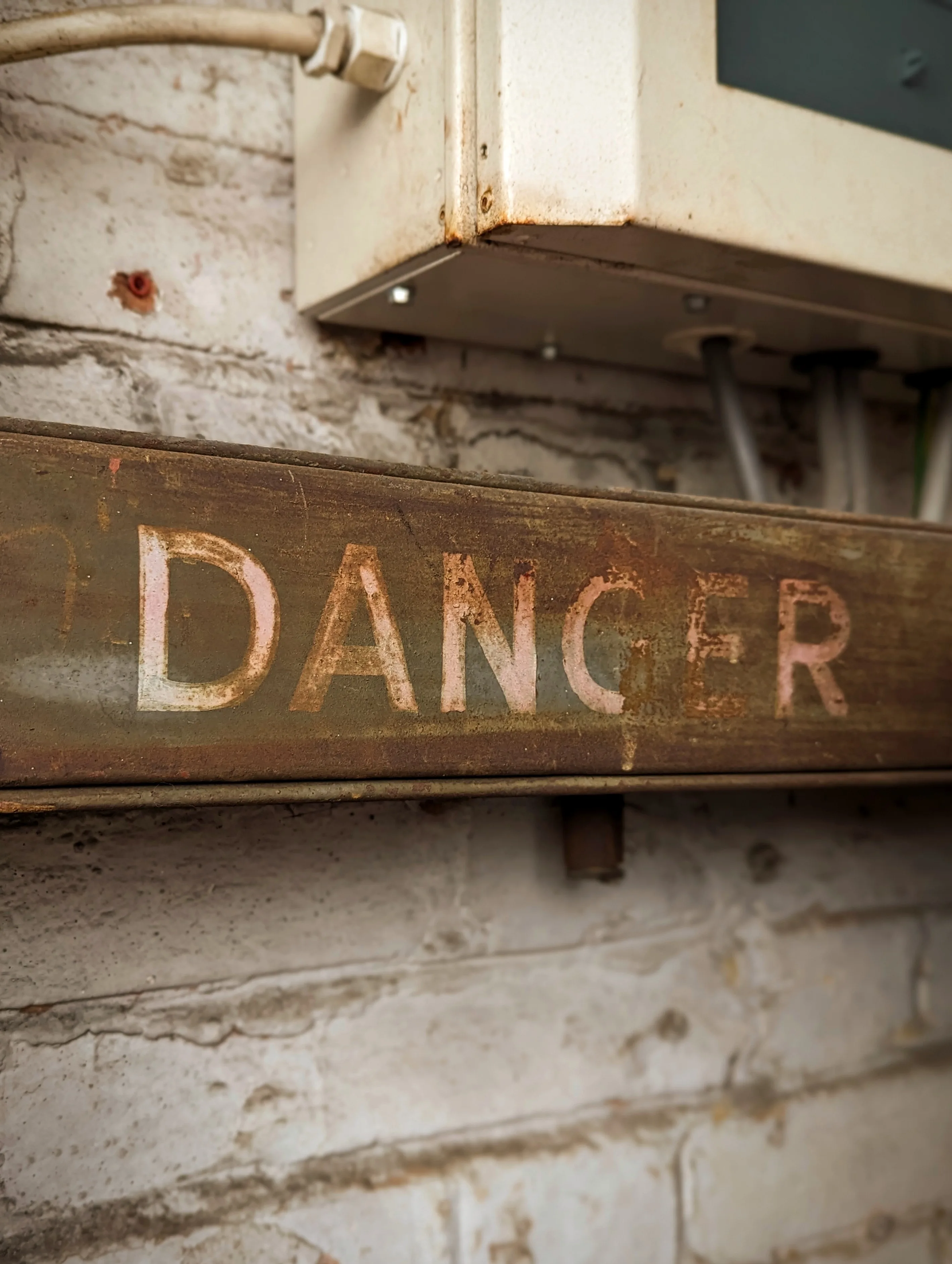 The word DANGER stenciled in fading pink letters on a rusty metal beam against a weathered white brick wall.