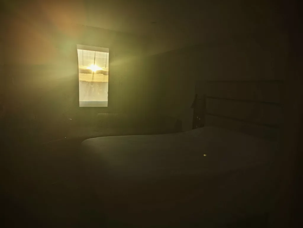 Golden sunlight streaming through a window into a dark bedroom, creating a warm lens flare that illuminates the corner of a bed.