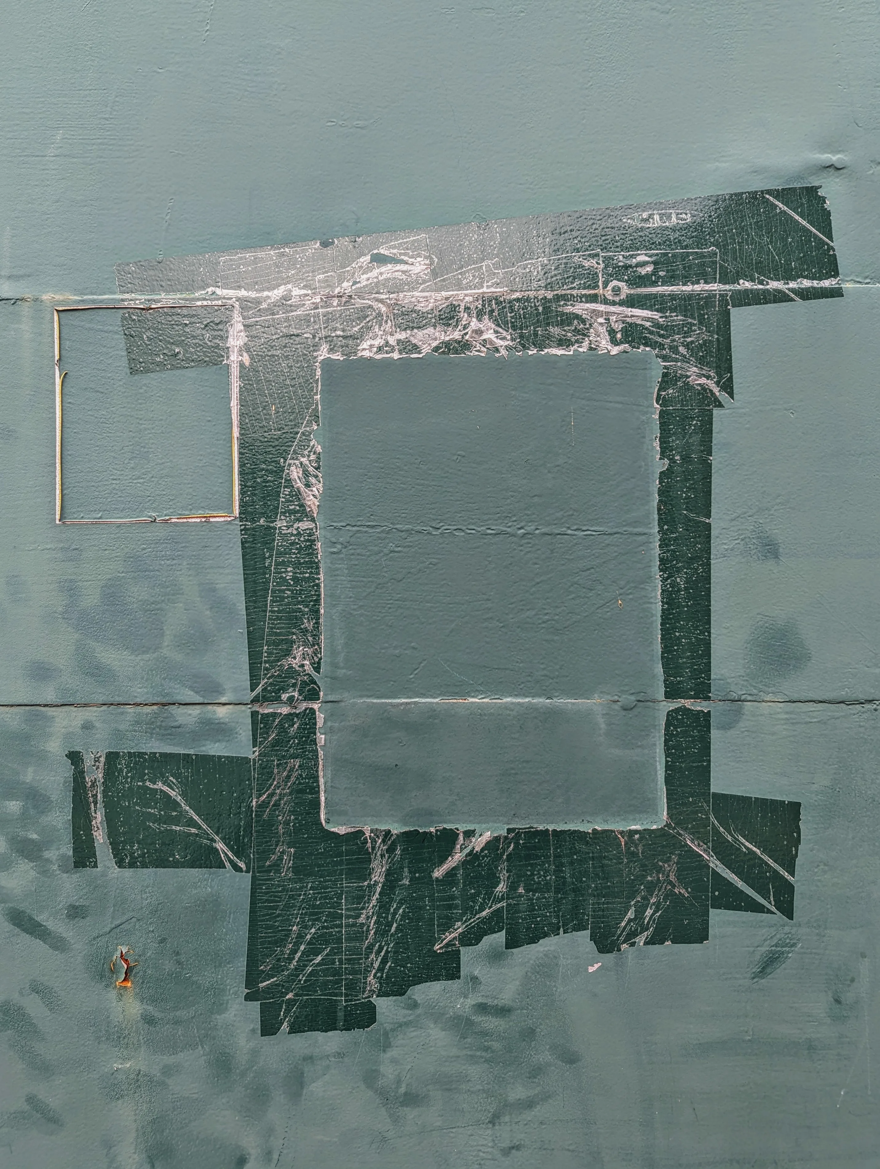Weathered dark green tape remnants forming a rectangular frame on a blue-grey painted metal wall beside a thin wire square outline.