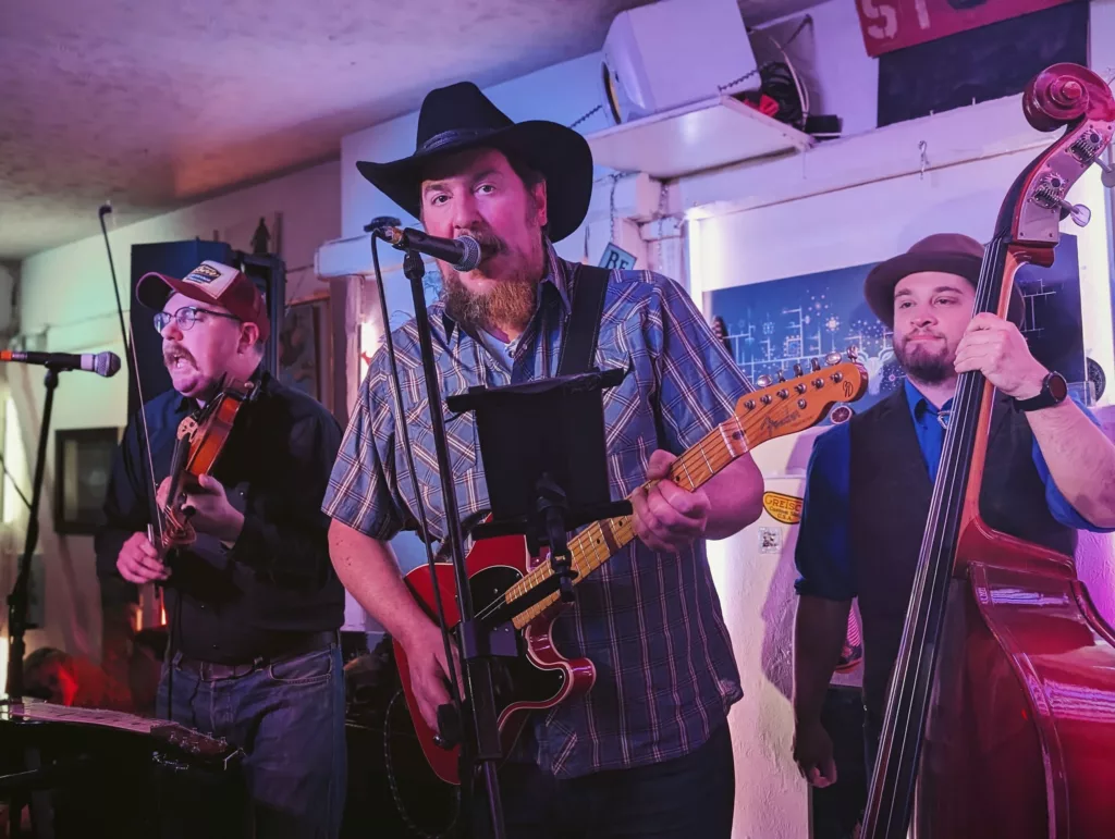 A country band performing under purple lights with a bearded singer in a black cowboy hat, a violinist, and an upright bassist.
