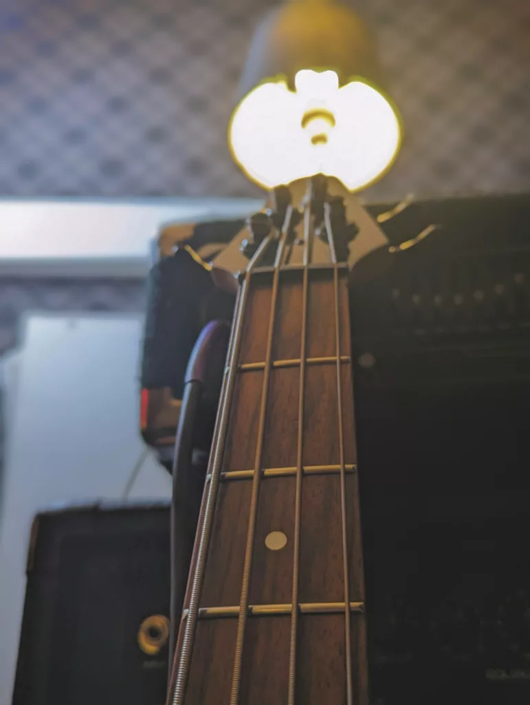Close-up looking up the neck of a bass guitar toward a bright circular light behind the headstock in a studio.