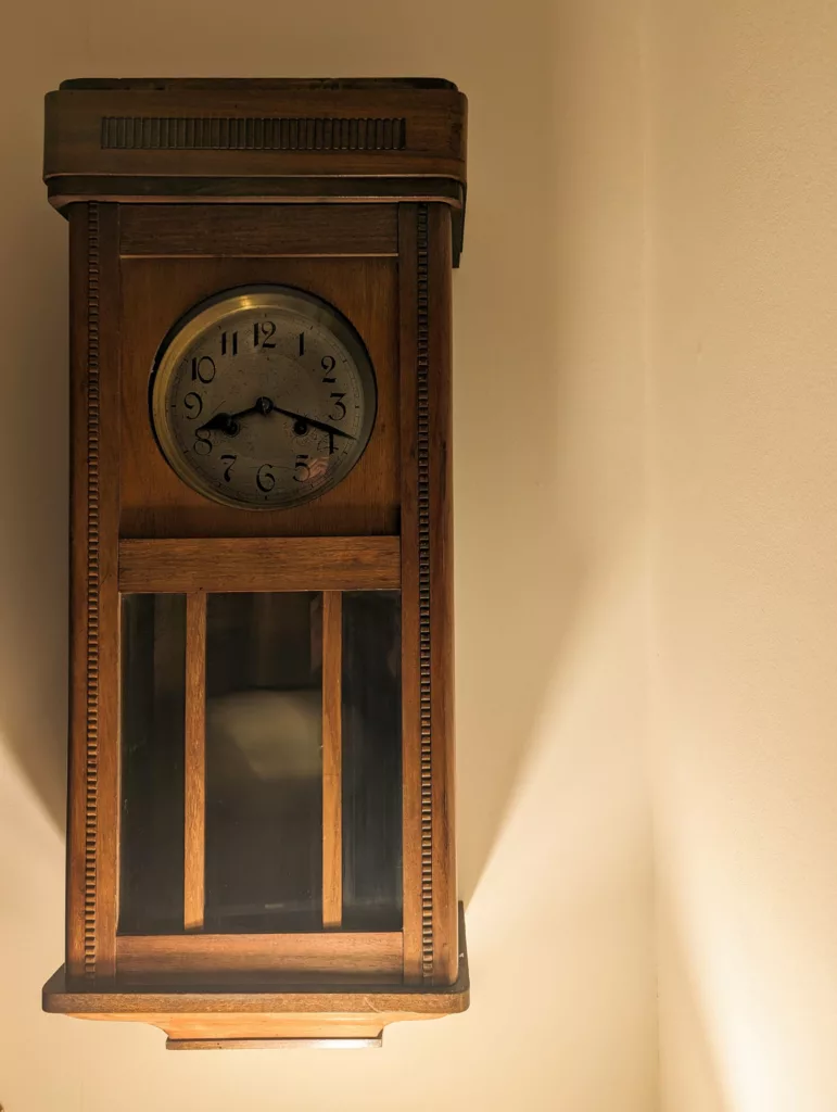 An antique wooden wall clock with a circular face and vertical slats over the glass pendulum window, mounted on a plain wall.