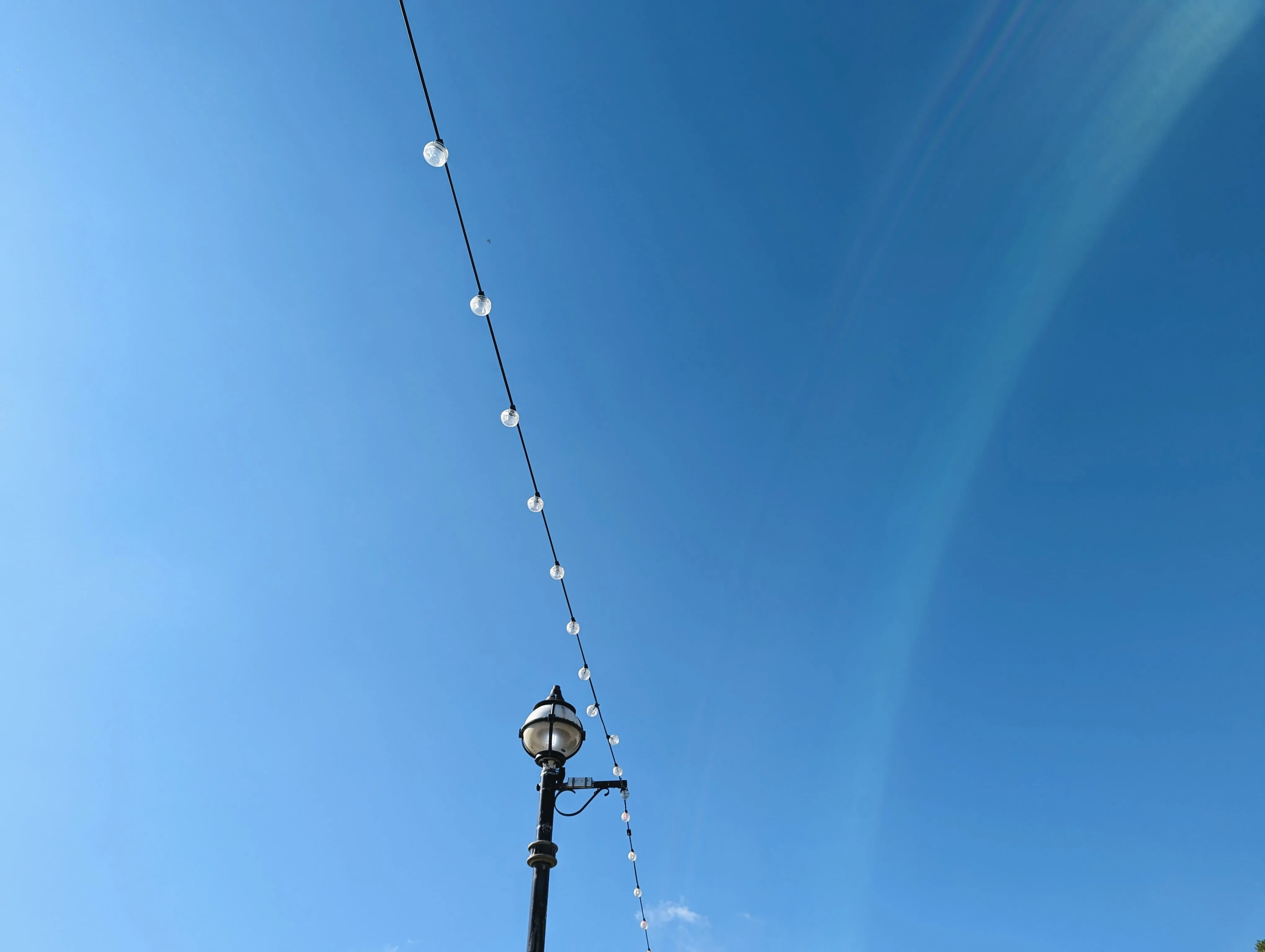 A diagonal string of festoon lights above a traditional globe-topped lamp post against a bright, clear blue sky.