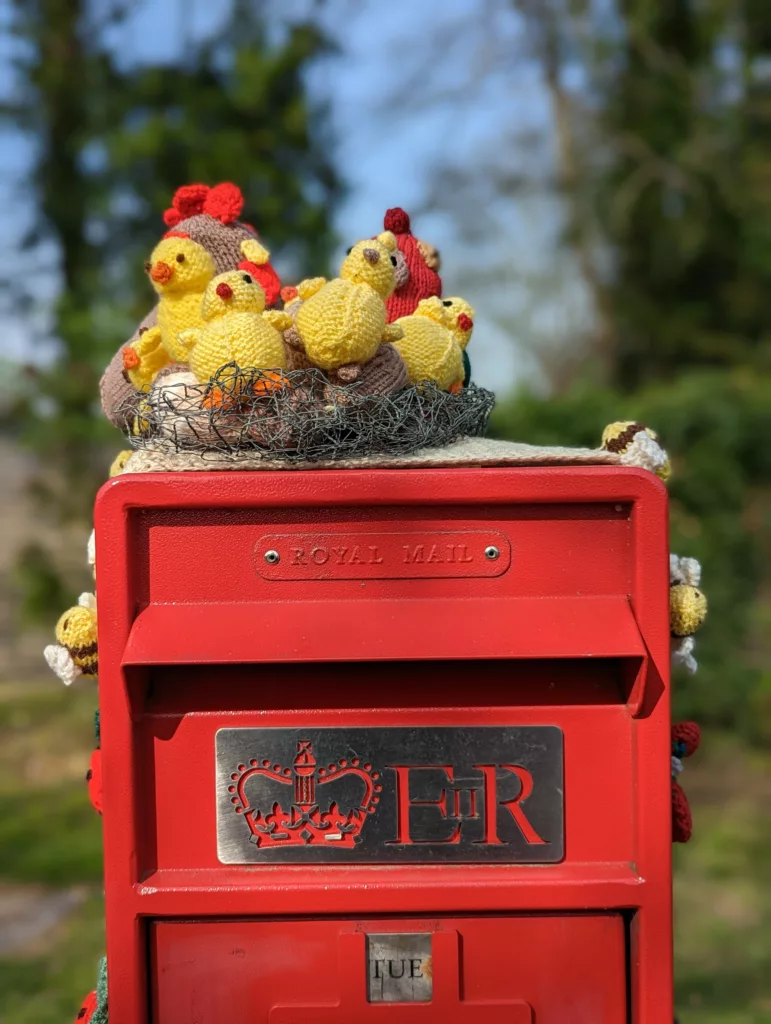 Knitted yellow chicks in a wire nest atop a red Royal Mail post box with the ERII insignia, set against a blurred green background.