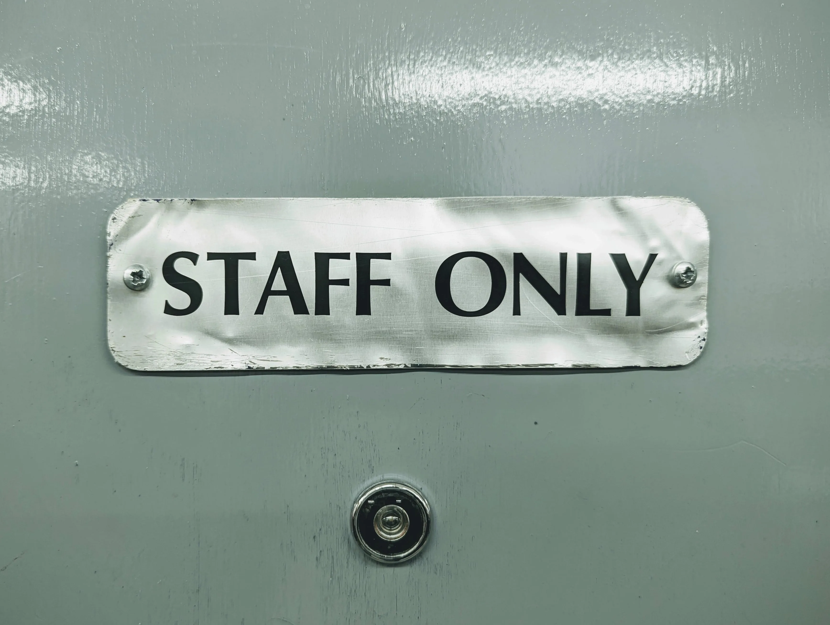 A brushed metal plate with the words STAFF ONLY screwed onto a grey painted door above a small peephole.