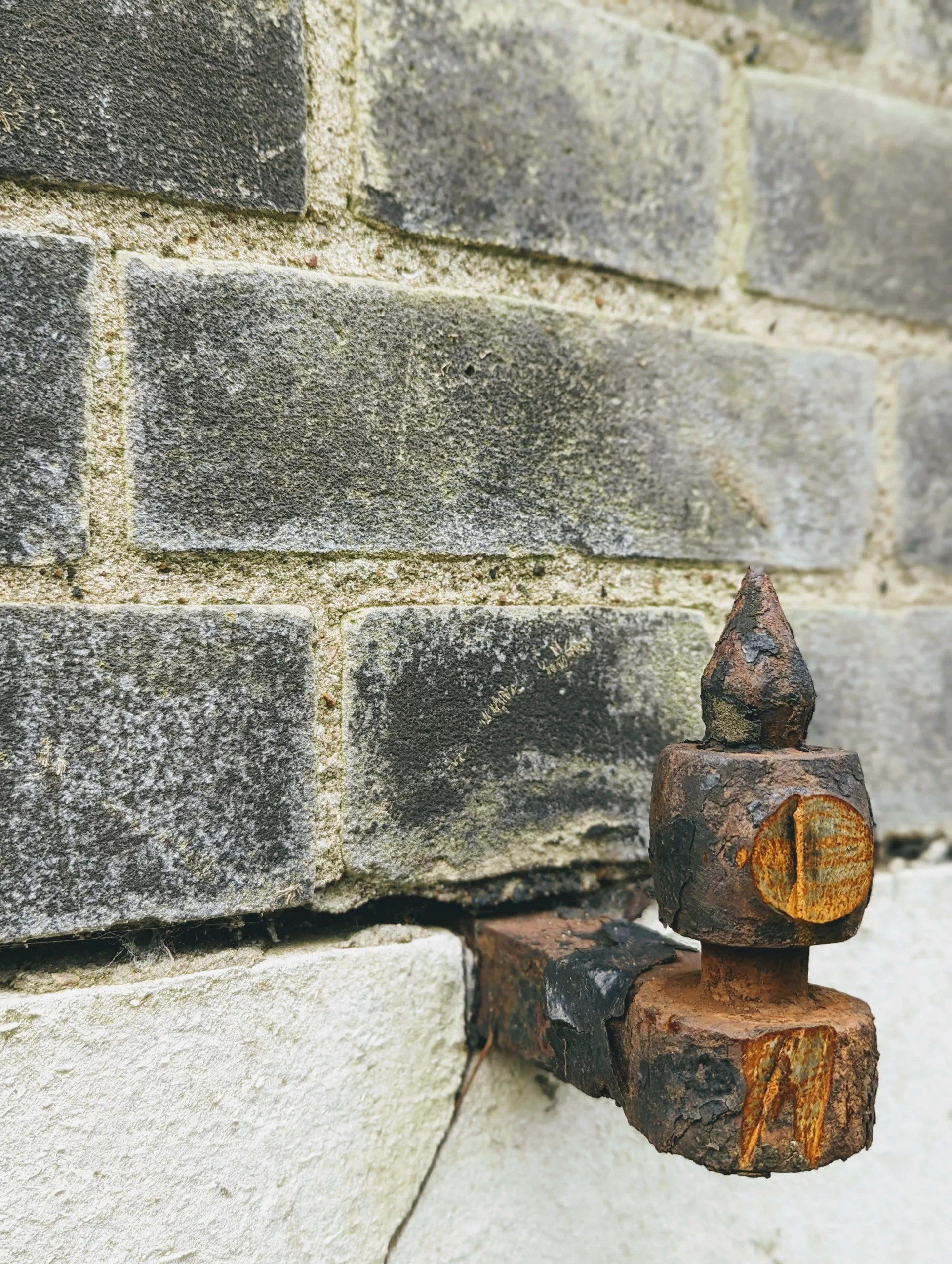 Rusted ornate metal gate hinge with a pointed finial protruding from a weathered grey brick wall above a white base.