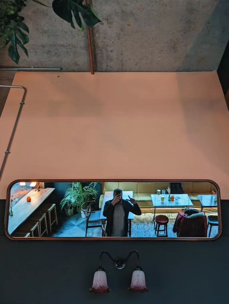Reflection of a person making a peace sign in a long mirror below a large peach wall and concrete ceiling in a cafe.