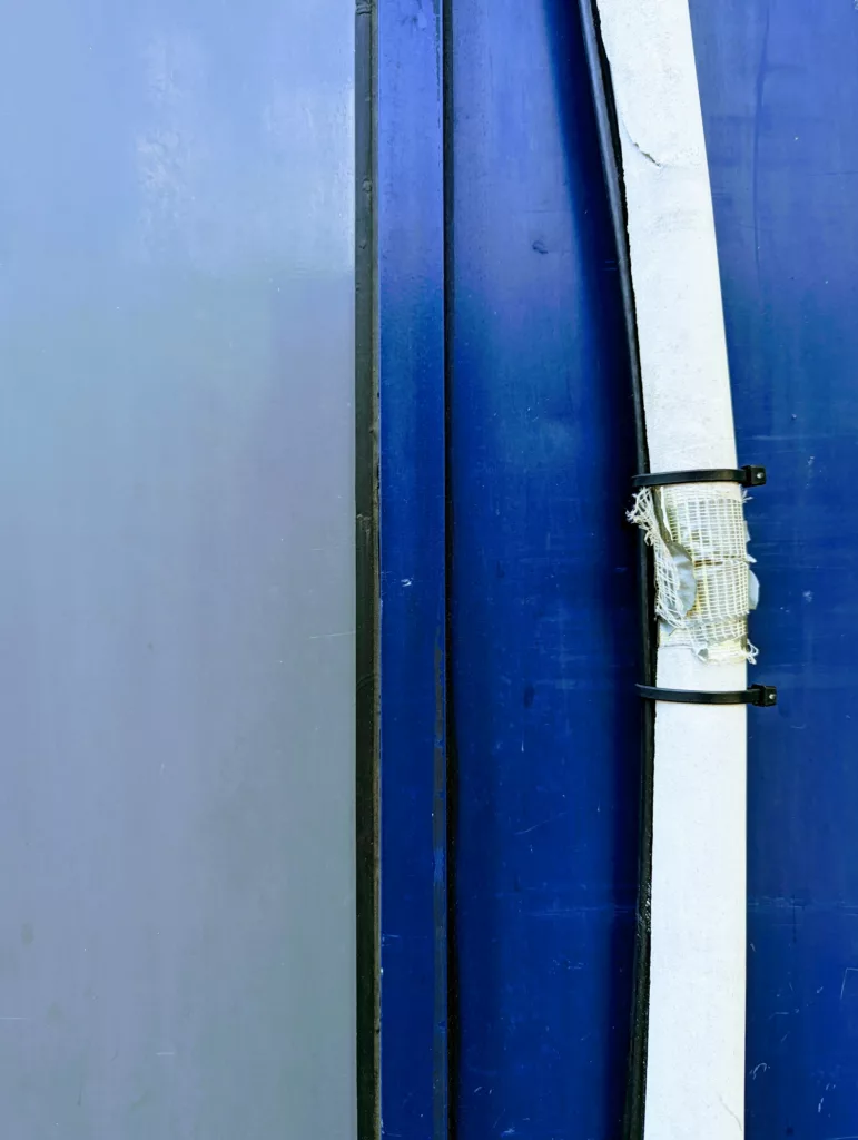 A white padded pipe with frayed mesh secured by black cable ties against a background of vertical light and dark blue panels.