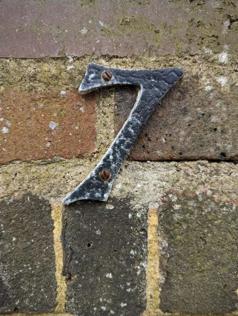 Weathered black metal house number seven screwed into a rustic brick wall.