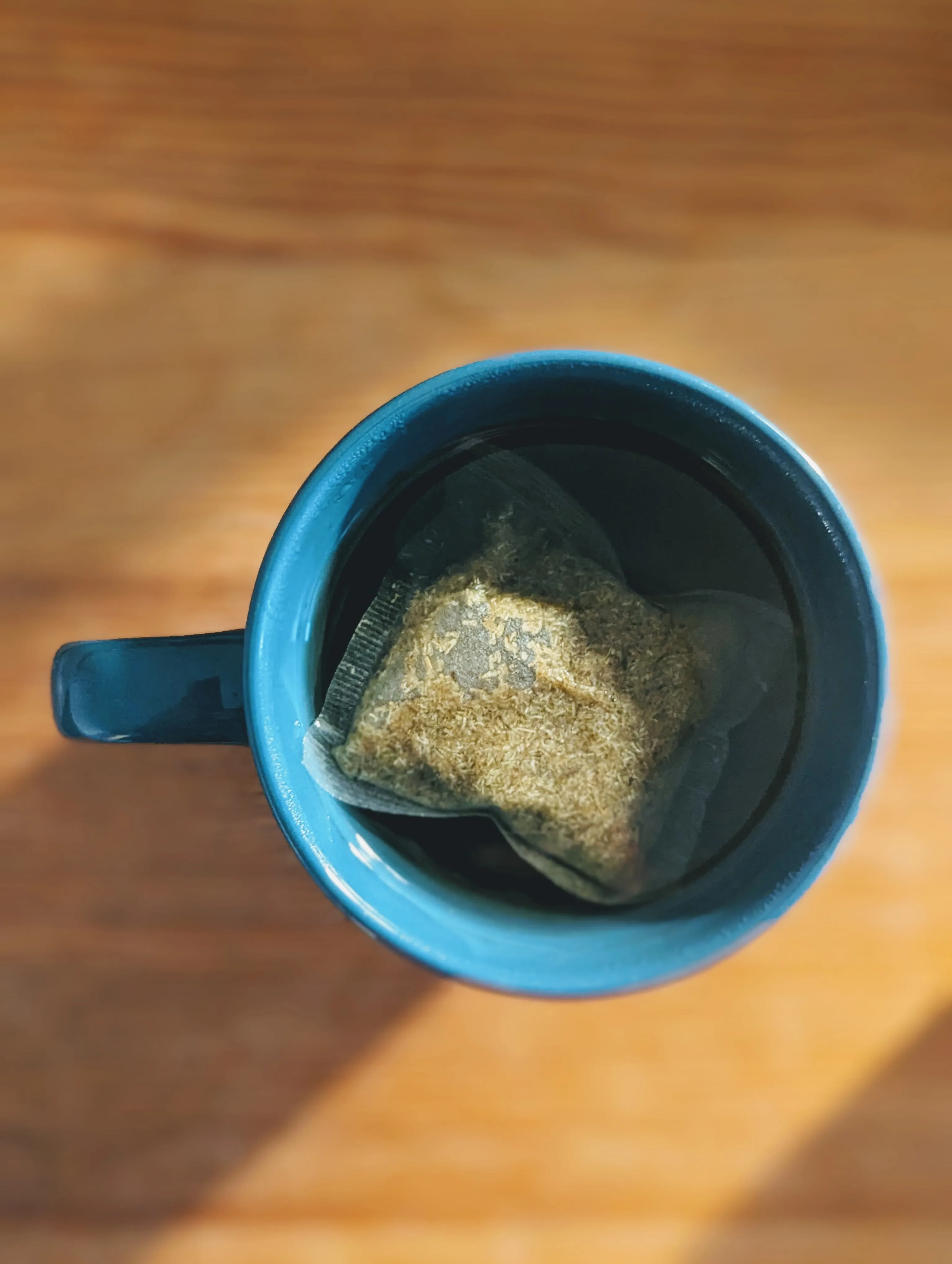 Top-down view of a herbal tea bag steeping in a blue ceramic mug on a wooden surface in soft sunlight.