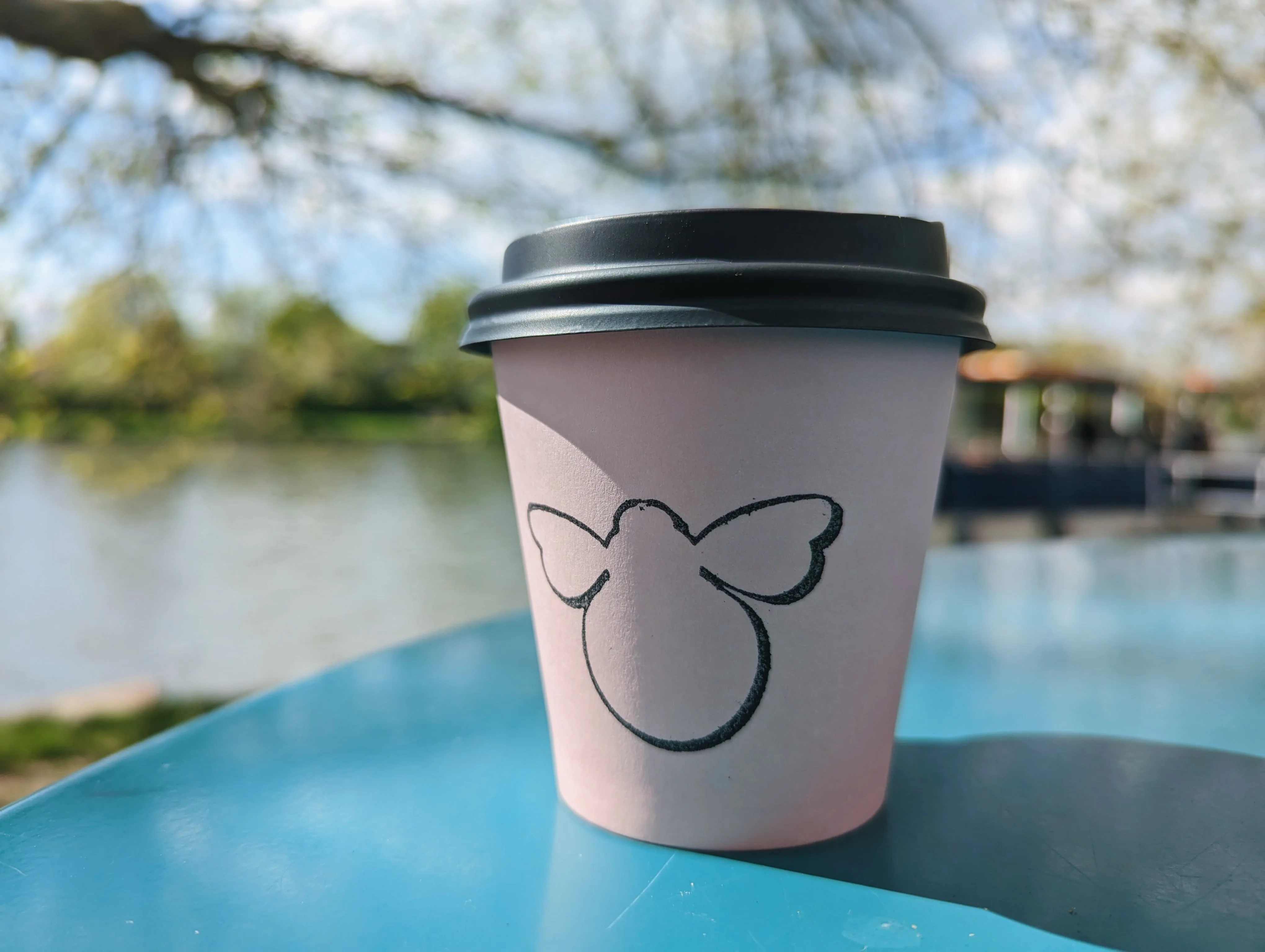 Pink paper coffee cup with a bee logo on a blue outdoor table with a blurred river and trees in the background.