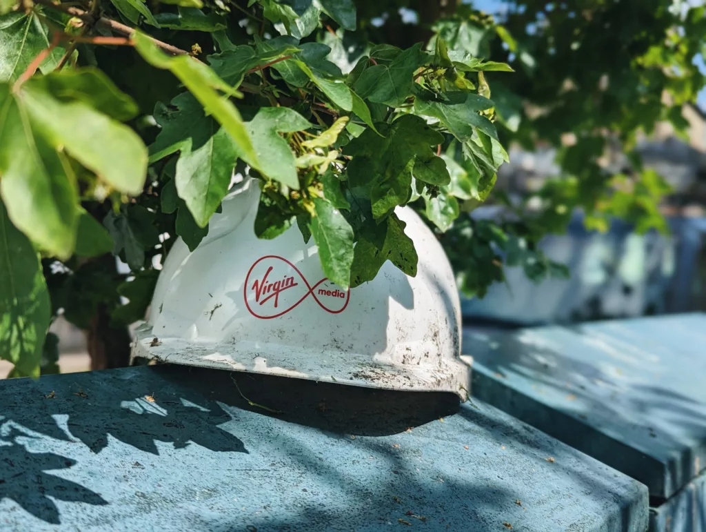 A white Virgin Media hard hat sits on a blue metal utility box, partially obscured by green leaves from an overhanging branch.