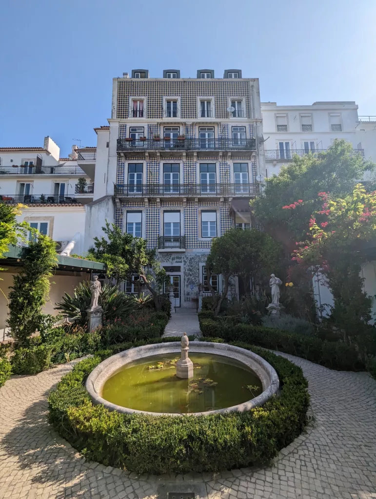 A round stone fountain in a manicured garden facing a tall building with a blue and white patterned tile facade and wrought iron balconies.